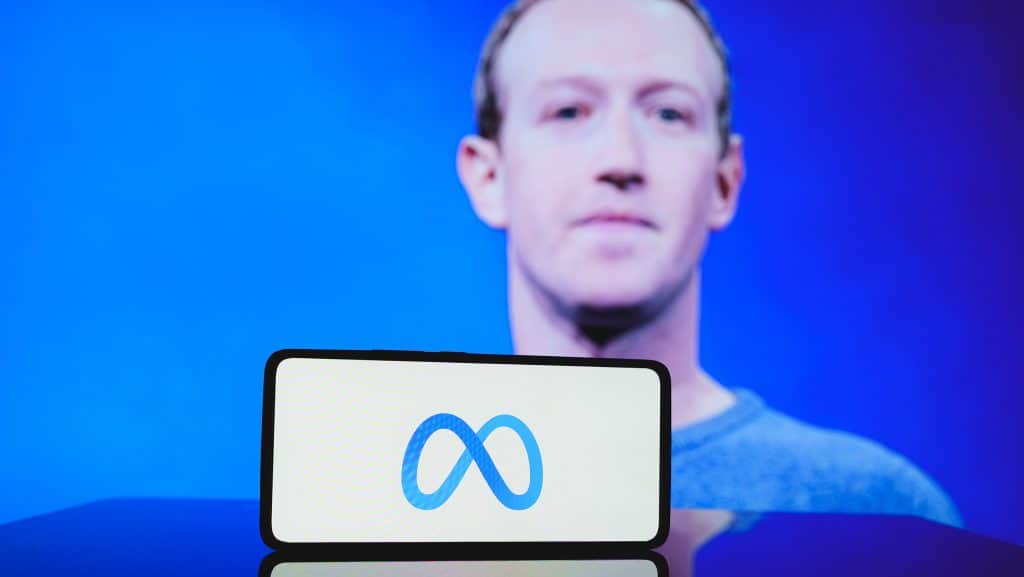 Ao fundo, foto desfocada de Mark Zuckerberg; à frente, logo da Meta em um smartphone